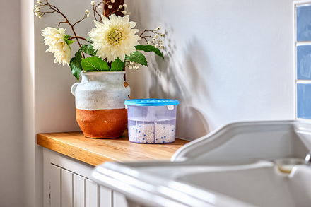 Zero In Air Ace Refillable Dehumidifier 300g moisture absorber with blue lid, positioned on a wooden kitchen worktop beside a vase of flowers to help manage excess moisture, condensation and damp.