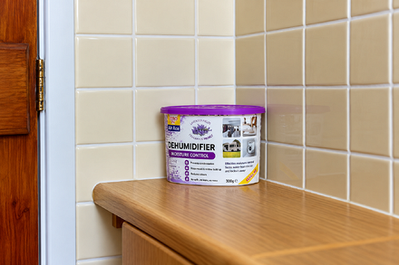 Zero In Air Ace Lavender Refillable Dehumidifier 300g with purple lid, positioned on a wooden countertop against cream bathroom tiles to help absorb excess moisture, reduce condensation and control musty odours.