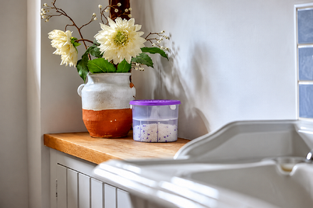 Zero In Air Ace Lavender Refillable Dehumidifier 300g with purple lid, positioned on a wooden kitchen worktop beside a vase of white flowers and a sink to help absorb excess moisture and reduce condensation and musty odours.
