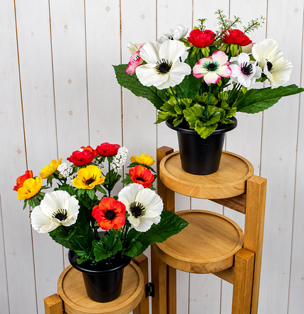 Two artificial anemone grave flower arrangements in black pots, featuring red, white, pink, yellow and orange blooms with green foliage, displayed on wooden stands.