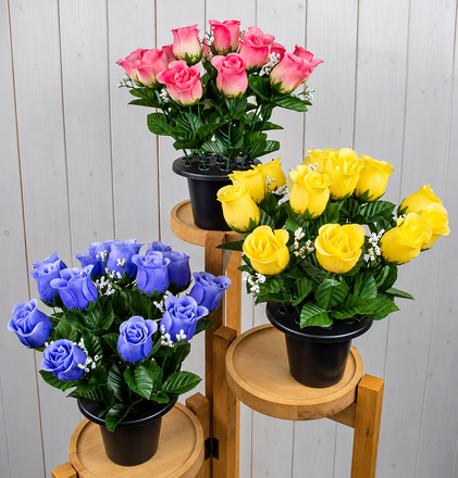 Three artificial rosebud grave pots featuring pink, yellow and purple-blue roses with green foliage and delicate white filler flowers, displayed in black pots on tiered wooden stands.
