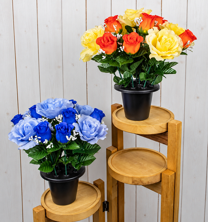 Two artificial rose and rosebud grave pots featuring yellow and orange roses in one arrangement and blue roses in the other, with green foliage and black pots displayed on wooden stands.